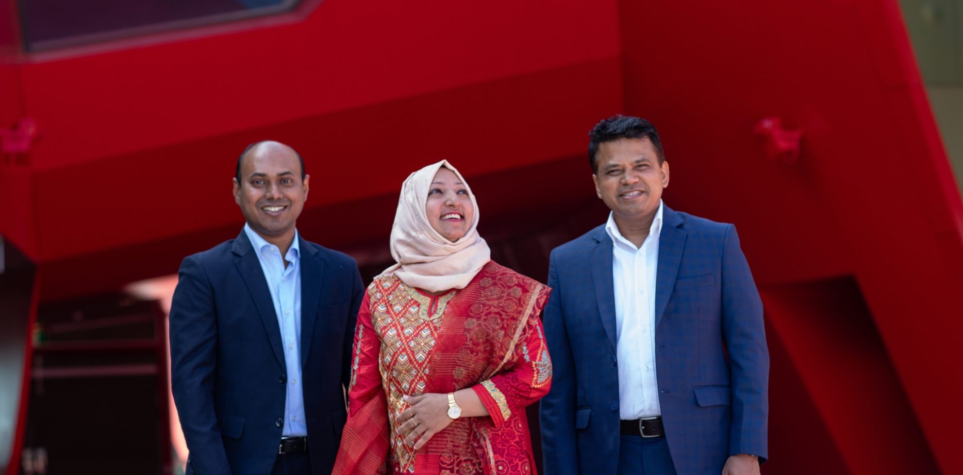 Three Australia Awards scholars standing side by side in professional attire against a red and dark blue background.